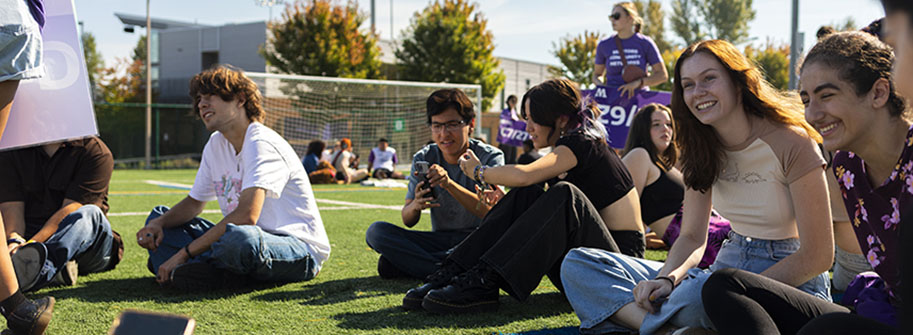 diverse group of seven students smiling and sitting on a lawn at convocation event