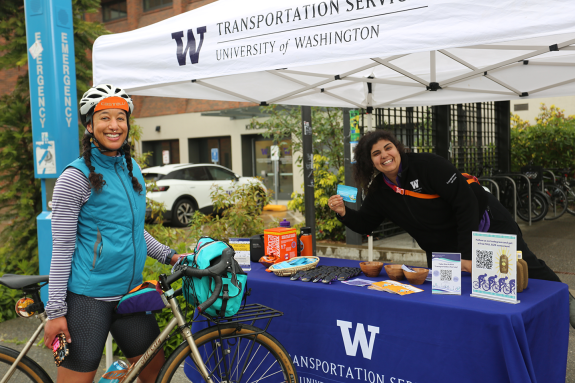 A smiling person in a blue vest standing next to and holding their white bicycle, at a transportation services table where a smiling employee looks at the viewer