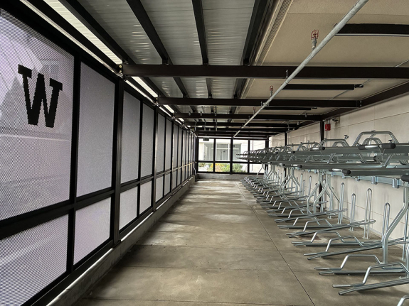 A view from inside the bike house, with double-decker bicycle racks running down a solid wall of white, and opposite them is a translucent wall of metal mesh with a W visible