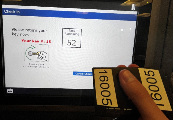 a person's hand holding a key fob in front of touch screen reading please return your key with key slot 15 listed